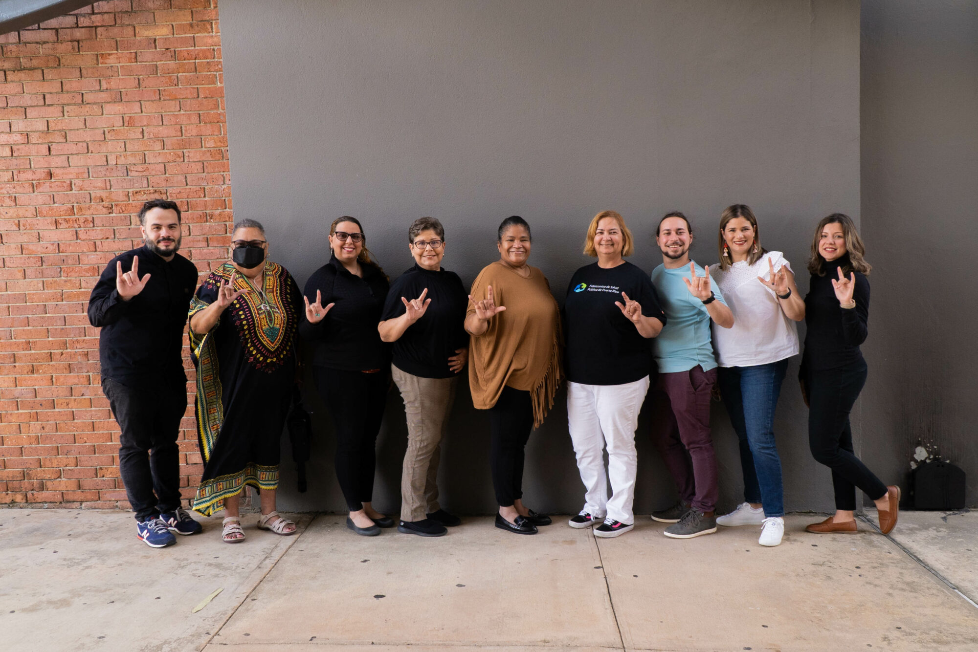Picture of 9 people from the Aqui Nos Cuidamos team and community leaders standing in from of a wall and doing the "I love you" sign in American Sign language.