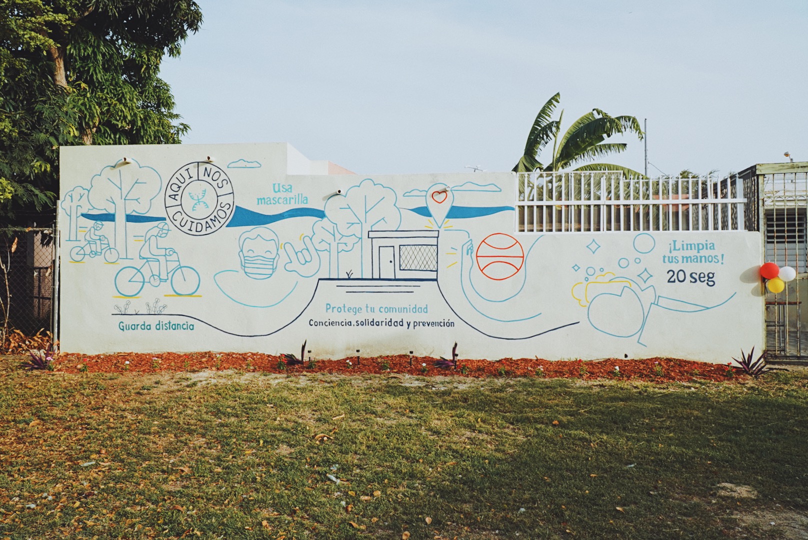 Picture of a Mural in a park with the logo of Aqui Nos cuidamos and that says in Spanish: Protege tu comunidad (protect your community) and with picture of handwashing and mask wearing.