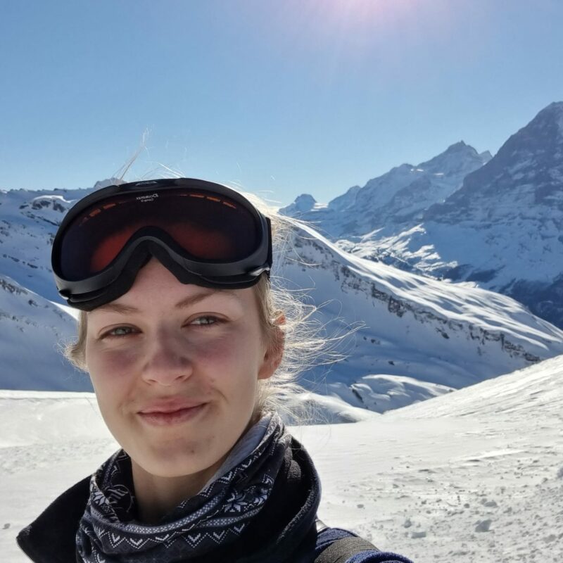 A photo of a woman wearing skiing goggles with mountains in the background.